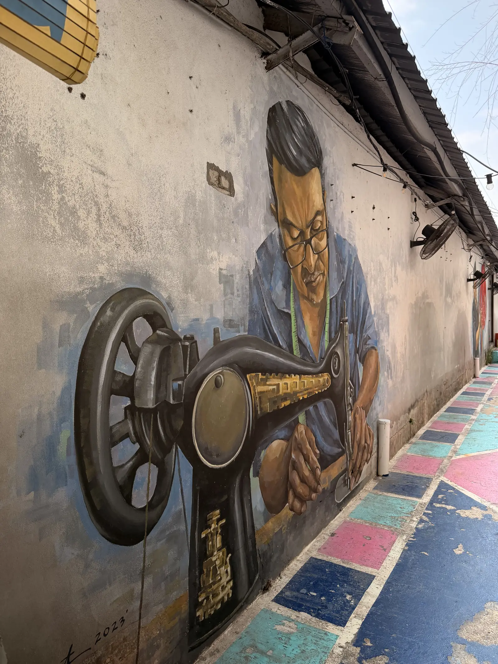 Wandbild eines Mannes an einer Nähmaschine in einer bunt bemalten Gasse