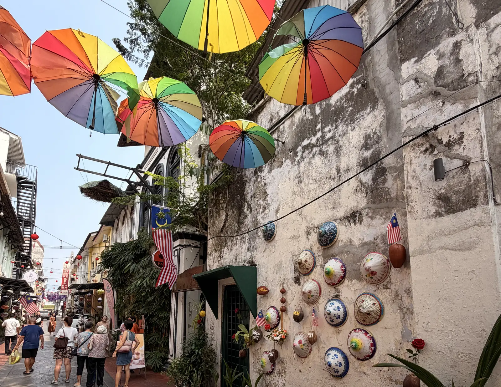 Bunte Regenschirme über der Market Lane in der Altstadt von Ipoh, an der Wand hängen Strohhüte